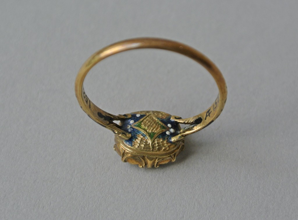 Mourning ring; gold; bezel containing rock crystal (or glass), beneath which is a monogram within looped border of gold thread over plaited hair; underside of bezel has lozenge enamelled in green upon ground of dark blue with white spots; enamelled on shoulders with blue lozenge with central white spot on ground of black enamel; each shoulder branches into three. No maker's mark.