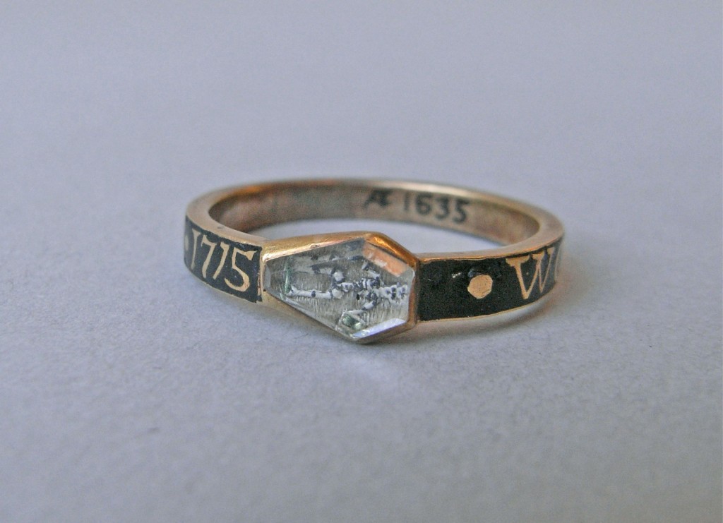 Mourning ring; gold; hoop with inscription reserved in black enamel; small coffin-shaped bezel with rock crystal over black and white enamelled skeleton holding arrow and hour-glass, upon background of hair. Green enamel on hour-glass and patch of ground on which skeleton stands. No maker's mark.