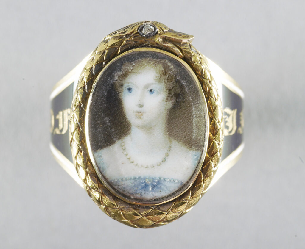 Mourning ring mounted with glazed oval miniature of Princess Charlotte (1796-1817) within a frame of a gold ouroboros with a diamond eye, the band enamelled in black and white with a mourning inscription 'IN MEMORY OF PRINCESS CHARLOTTE 1817'. This miniature, which in terms of technique is consistent with Jones's work, is based on a miniature by her dating from 1813. The primary evrsion, in the Palais Royal. Brussels, shows the Princess, standing, with Westminster Abbey in the background on the left and was used as a basis of a stipple engraving by John Agar. Another version is at Anglesey Abbey. This miniature shows the Princess wearing a pearl necklace rather than the more elaborate emerald and pearl necklace in the source.