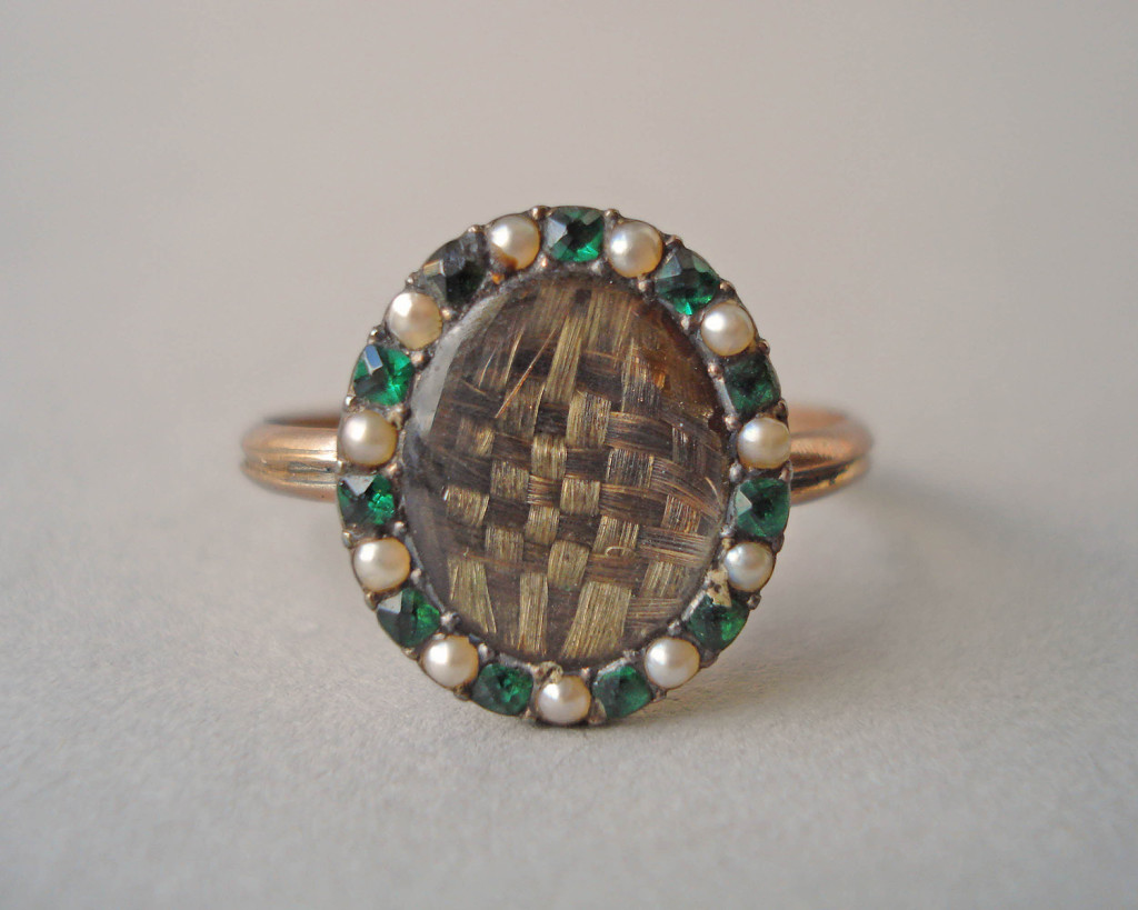 Mourning ring; gold; slender channelled hoop; oval bezel containing plaited hair covered with convex glass; border of seed pearls alternating with emerald pastes. No maker's mark.