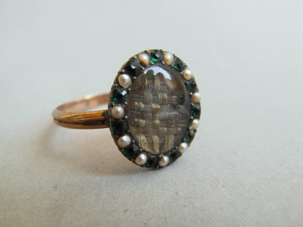 Mourning ring; gold; slender channelled hoop; oval bezel containing plaited hair covered with convex glass; border of seed pearls alternating with emerald pastes. No maker's mark.