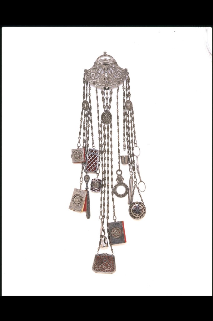 A chatelaine would have hung from a lady’s waist and was intended to be both decorative and practical. The small tools and accessories that might be incorporated included a watch, scissors, tweezers, magnifying glass, scent flask and miniature notebook or ivory writing tablet. Cut steel was a fashionable material for jewellery, buttons, buckles, sword hilts and watch chains in the decades around 1800. They were made from brightly polished rivets, their ends faceted to imitate diamonds. Such pieces gave a grey but powerful glitter. Originally an English speciality, the production of cut steel had spread to other centres in Europe by the early 19th century. The crowned monogram decorating the hook-plate has not been identified.