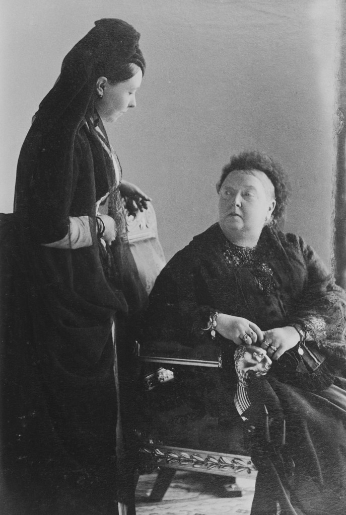 The Empress Frederick (left) stands, looking down at her mother, Queen Victoria, who is seated and looking up at her daughter. Both in mourning dress. 