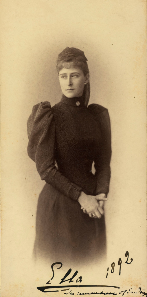 Photograph of Grand Duchess Elizabeth Feodorovna. She is standing with her head turned to the left and her hands clasped together. She is wearing dark mourning clothes with a cross at the collar. The photograph is annotated 'Ella 1892 In remembrance of Sandringham'. Grand Duchess Elizabeth Feodorovna, previously Princess Elisabeth of Hesse, married Grand Duke Sergei Alexandrovich in June 1884. In this photograph she is probably in mourning for her cousin Prince Albert Victor, Duke of Clarence and Avondale, who died at Sandringham in January 1892. 