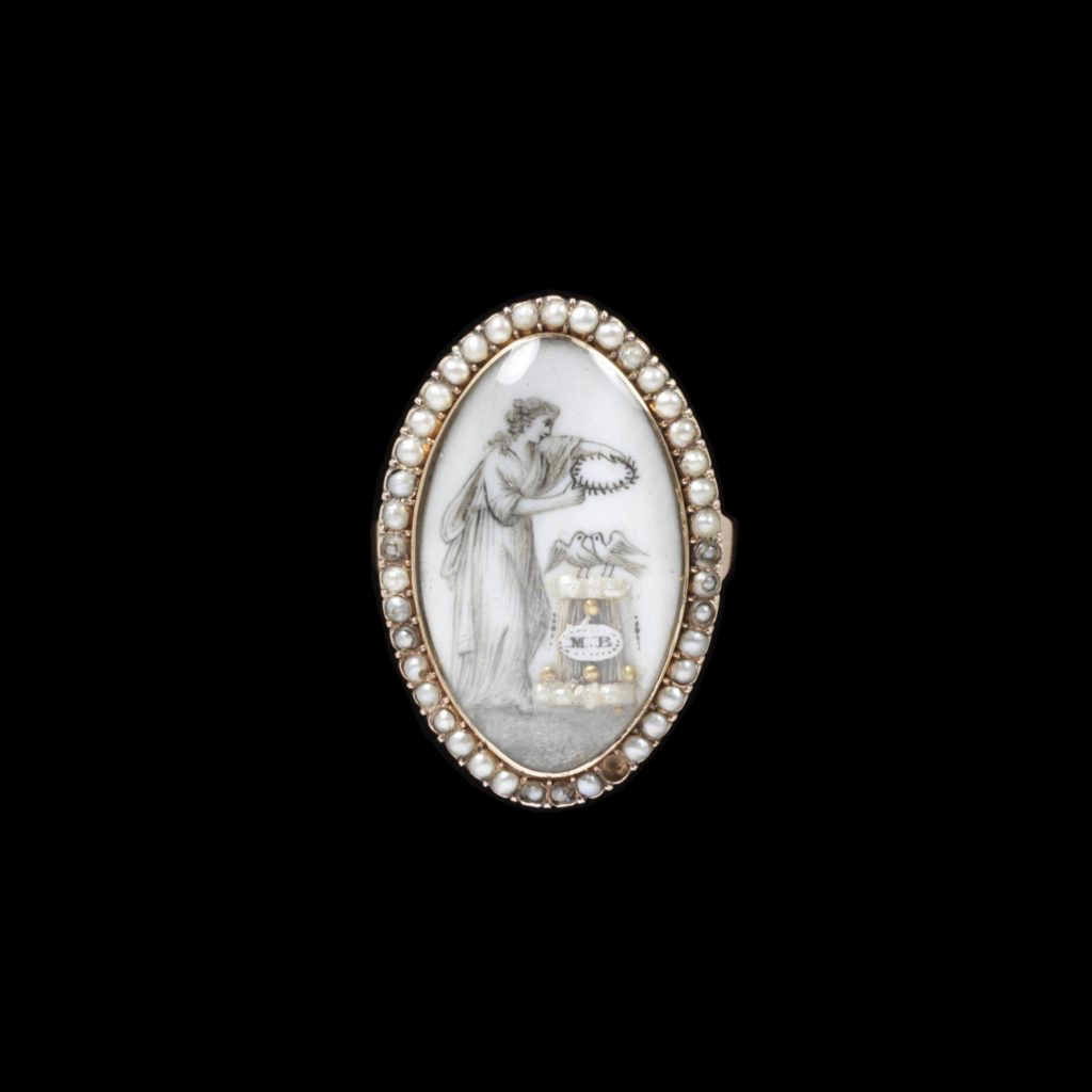 Gold ring set with pearls. A marquise bezel with a sepia painting under crystal of a woman holding a garland over a pair of doves perched on an altar bearing the initials 'MB', executed in hairwork and decorated with pearls and gold details, with a half-pearl border.