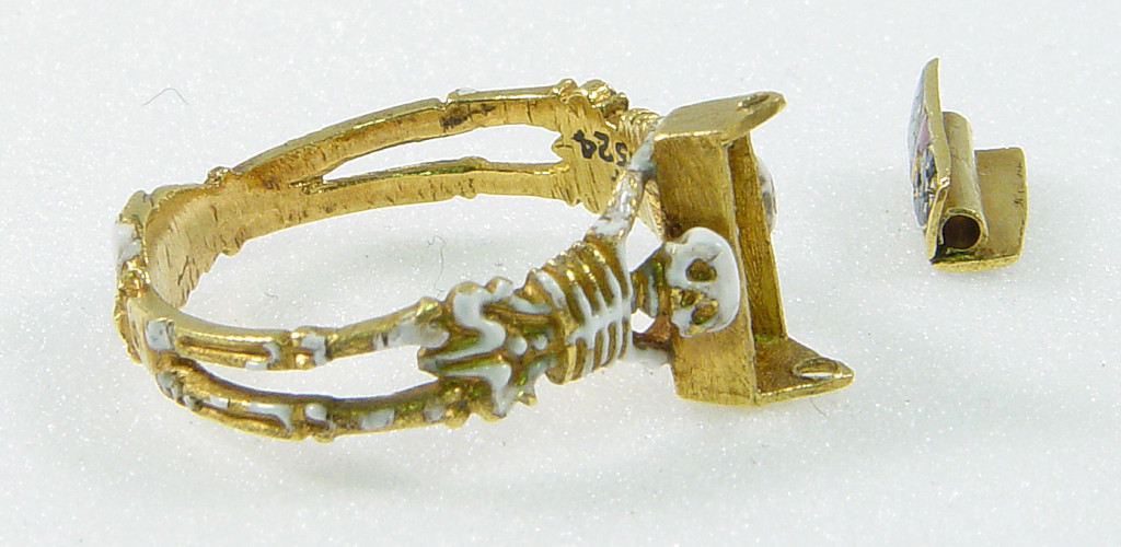 Mourning-ring; enamelled gold, the hoop enamelled white, in the form of two skeletons supporting a coffin-shaped bezel with moveable lid fastened with a pin and ornamented with a Maltese cross once enamelled red, on a black ground with hearts reserved in the metal, an enamelled cinquefoil at one end. Inside the coffin is a tiny white-enamelled skeleton. At the back of the bezel ares two clasped hands.