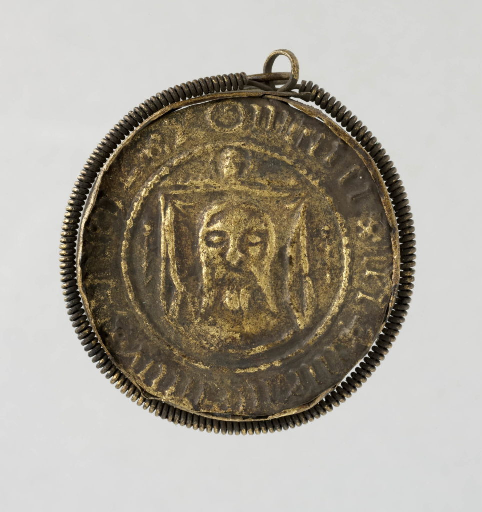 Pilgrim-badge; brass; circular; set in rim, round which is scroll of twisted wire and suspension loop; embossed with representation of St Veronica holding out the Vernicle