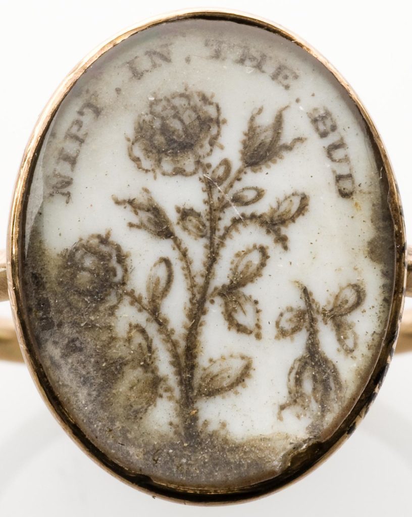 Gold memento mori ring, with oval setting which shows a flower and an inscription painted on a white background, beneath glass, possibly, glass, possibly 18th century. Detail view of flower and inscription on ring. White background. Wellcome collection at the Science Museum.