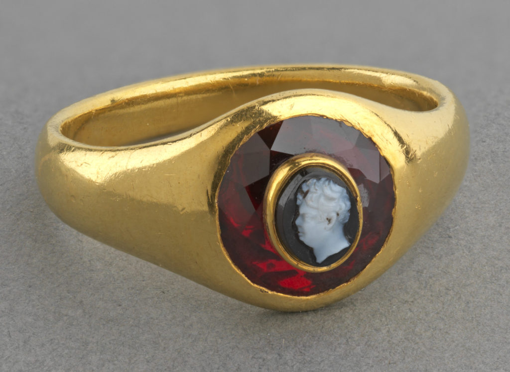 Plain gold ring. Faceted garnet inserted into the bezel and backed with foil, onyx cameo with left profile portrait of King George IV (1762-1830). Throughout the Regency and the 1820s the Royal goldsmiths, Rundell, Bridge & Rundell, regularly supplied George IV with finger rings set with cameos and intaglios cut with his likeness. These varied enormously in both price and design but most, it would seem, were intended as gifts to the King’s favourites. George IV presented the 6th Duke of Devonshire with a gold and enamel ring set with a cameo of the King on New Year’s Day, 1828. This ring, however, set with a miniature cameo of George IV inlaid into a garnet, appears to be without parallel. The cameo, the smallest in the collection, is finely cut with a detailed portrait. Although not identifiable in any of Rundell’s bills, the ring was probably supplied through them to George IV. Text adapted from Ancient and Modern Gems and Jewels in the Collection of Her Majesty The Queen, London, 2008