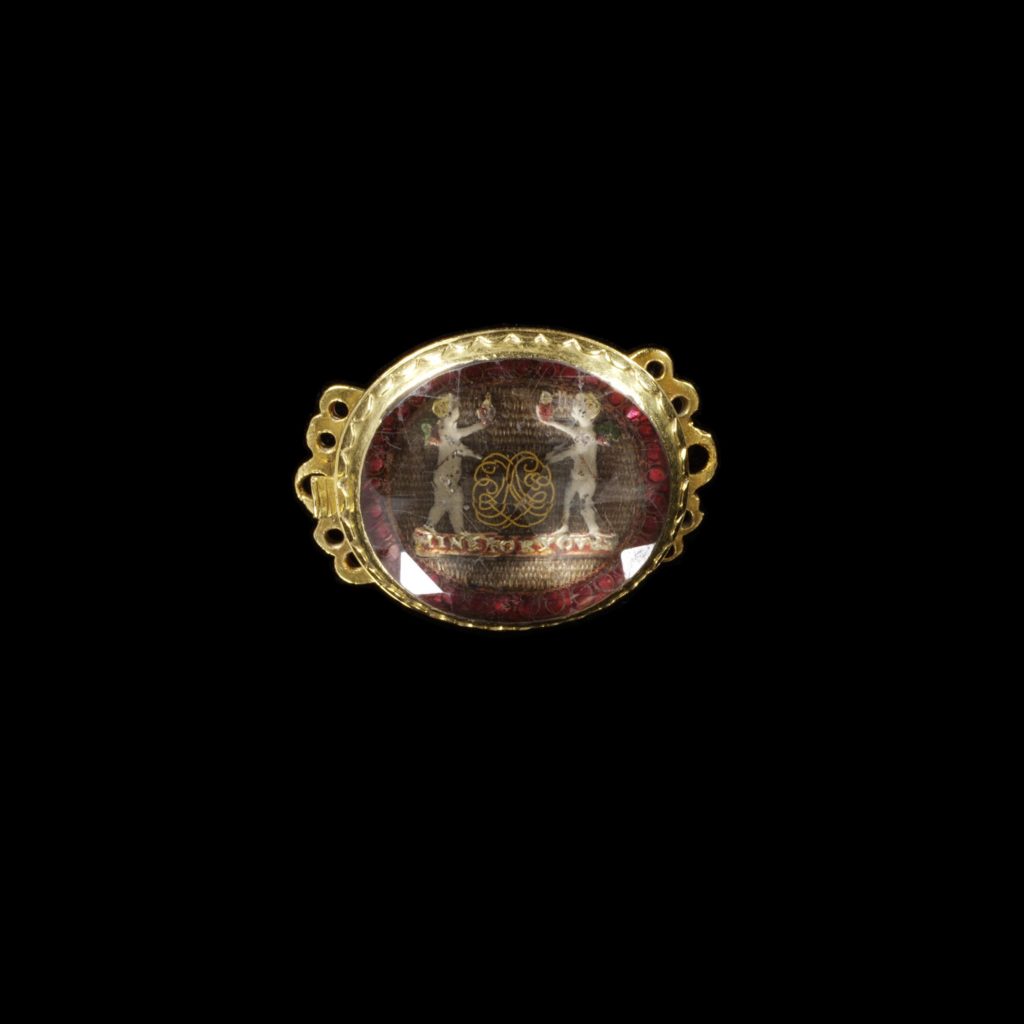 Gold slide-clasp, with a pair of enamelled Cupids holding flaming hearts with a gold wire cipher between, above the motto Mine for Yours on a background of hair and foil, under rock crystal, c.1700. V&A
