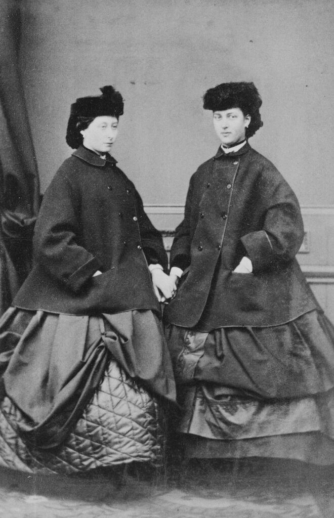 Alice, Princess Louis of Hesse and Princess Alexandra of Denmark, November 1862. They both wear overcoats and caps over large crinoline-supported dresses. Royal Collection UK. 