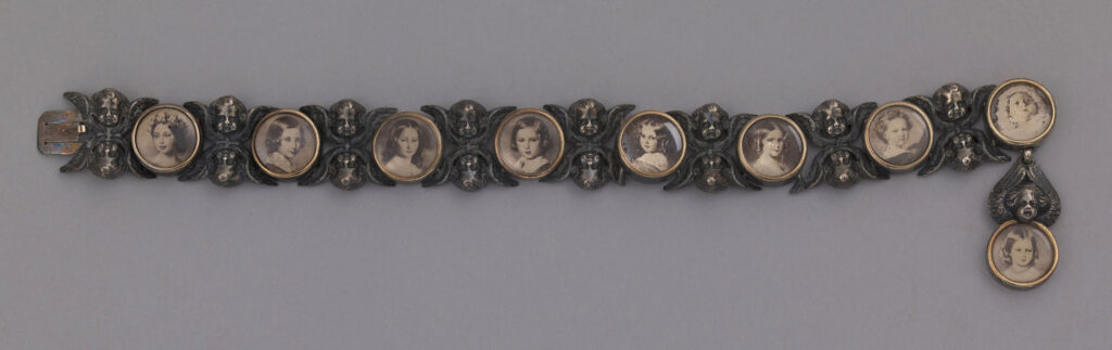 A silver-plated bracelet featuring nine photographic portraits of Queen Victoria and Prince Albert's children. The portraits are framed and joined by double-winged putti heads. The final portrait of Princess Beatrice is suspended by a single winged putto head. As the bracelet was given to Queen Victoria in 1854, it is likely the putto feature, holding a photographic portrait of Princess Beatrice, was added later, after her birth in 1857.